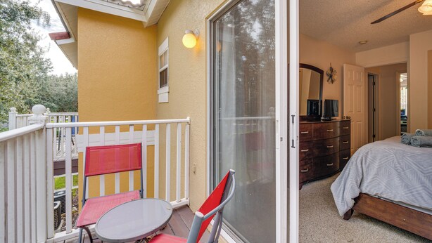 Un balcon avec une table et des chaises, une porte-fenêtre coulissante donnant sur une chambre à coucher équipée d’un lit et d’une commode.