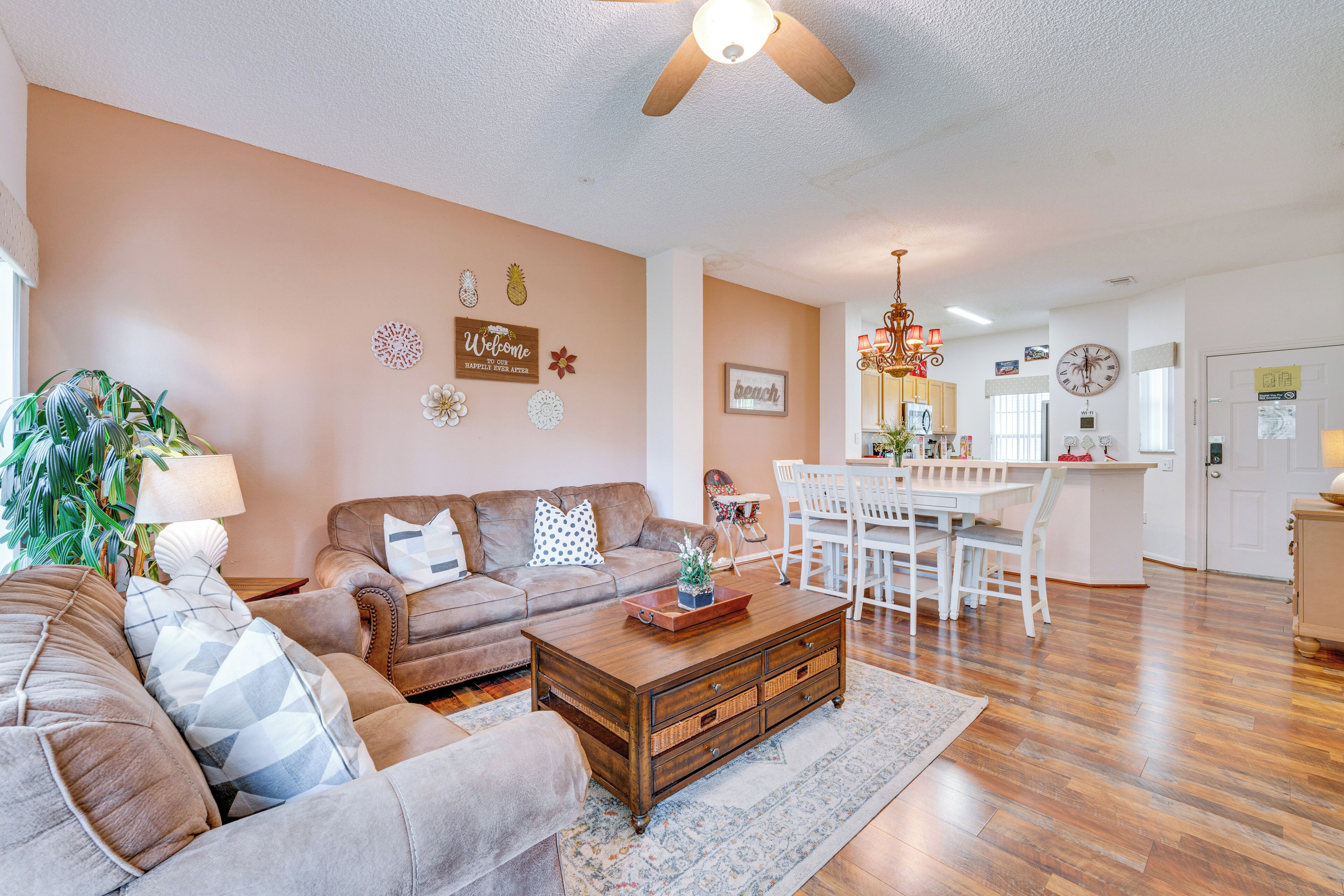 A living room with a sofa, coffee table, and decorative items. There is a dining area with a table and chairs. The room has wooden flooring and a ceiling fan.