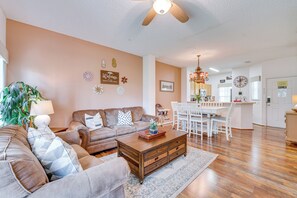 A living room with a sofa, coffee table, and decorative items. There is a dining area with a table and chairs. The room has wooden flooring and a ceiling fan.