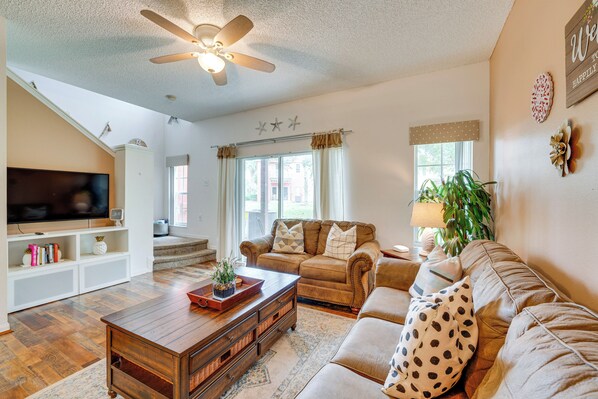 A spacious living room with a large flat-screen TV, a ceiling fan, and a staircase leading to an upper level.