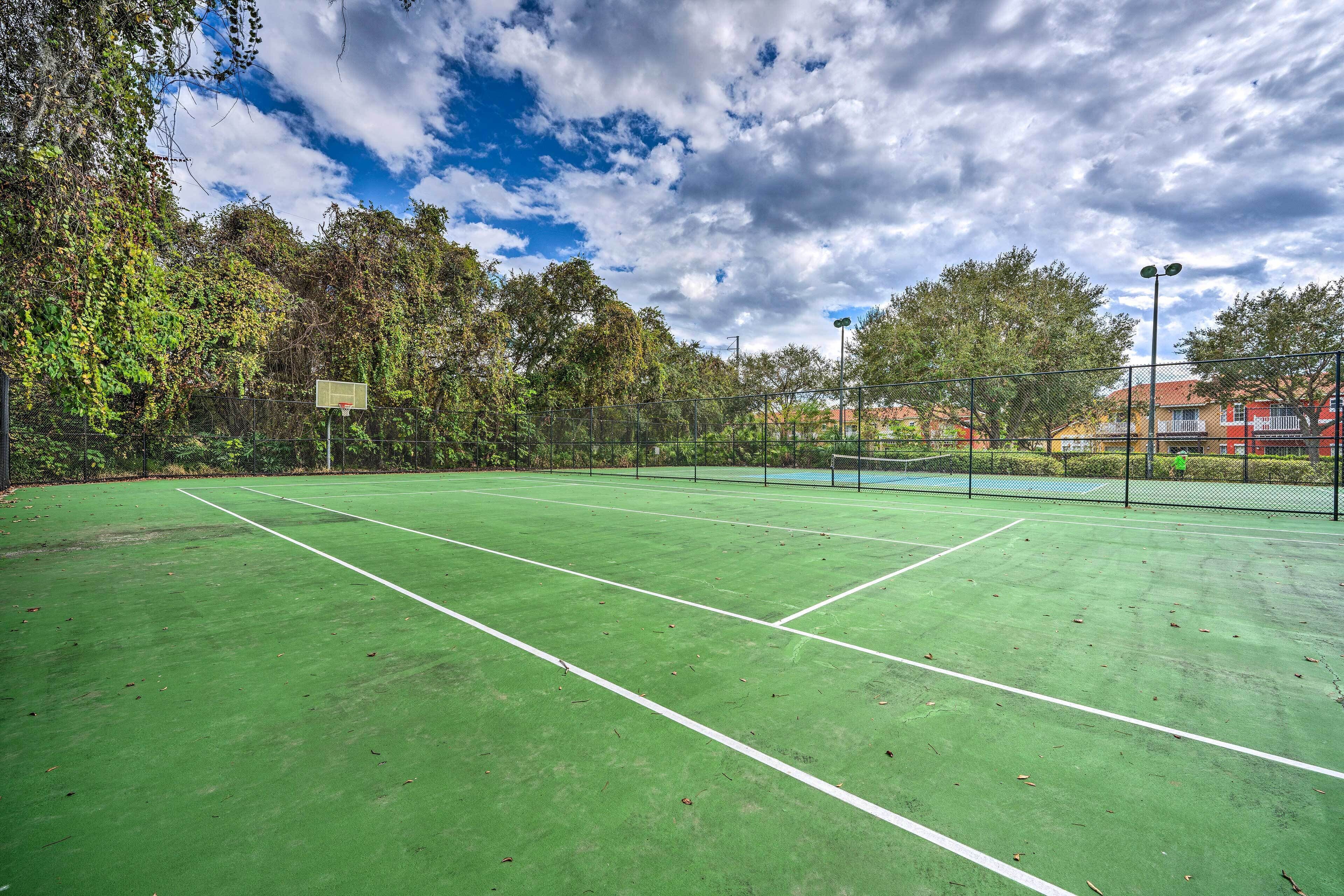 An outdoor tennis court with a green surface, surrounded by trees and a cloudy sky.