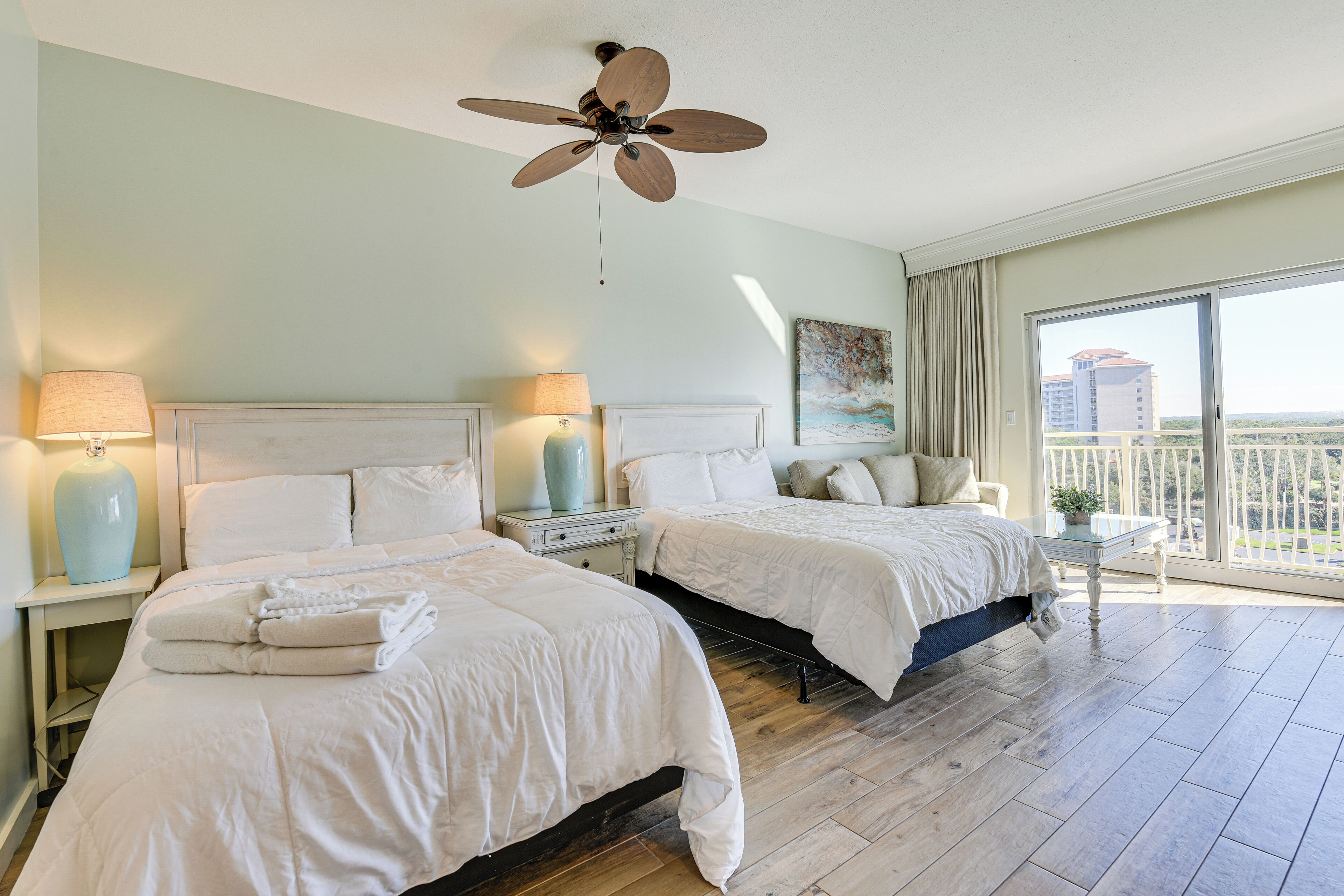 A hotel room with two beds, a ceiling fan, a large window with a view, and a nightstand with a lamp.