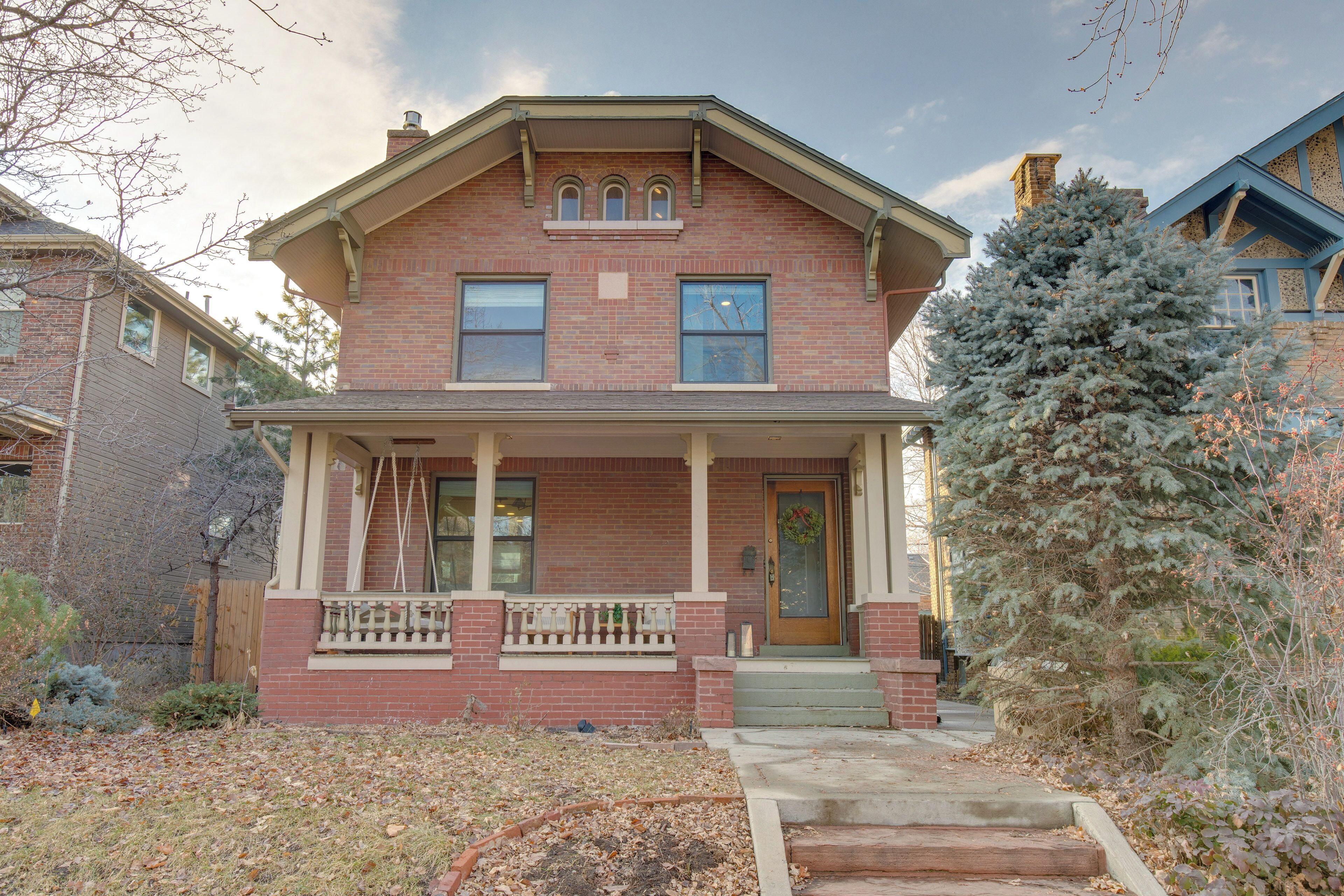 A two-story brick house with a front porch, a chimney, and a snow-covered evergreen tree.