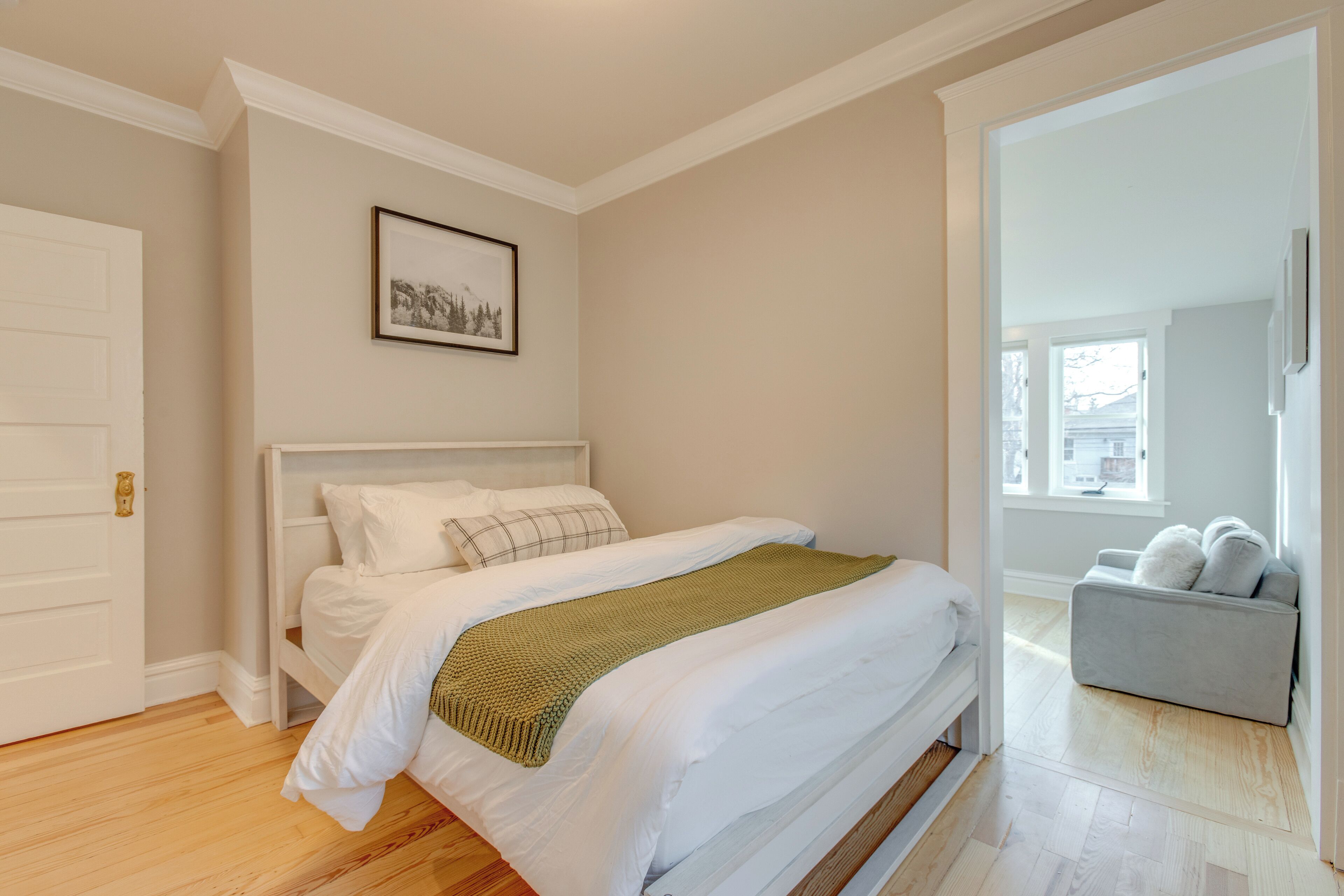 A bedroom with a bed, a framed picture on the wall, and a grey sofa in the adjacent room.