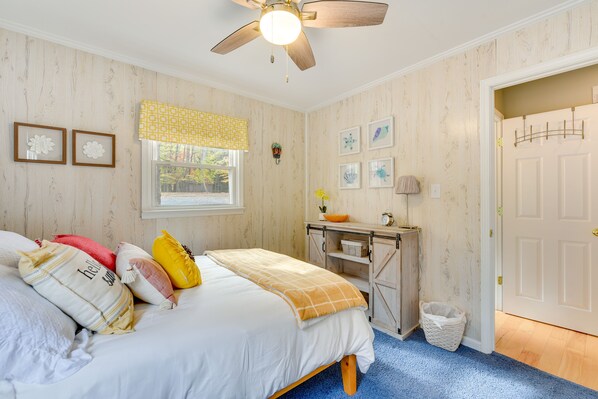 A bedroom with a bed, a ceiling fan, a window with a yellow valance, a desk with a basket, and a door with a coat rack.