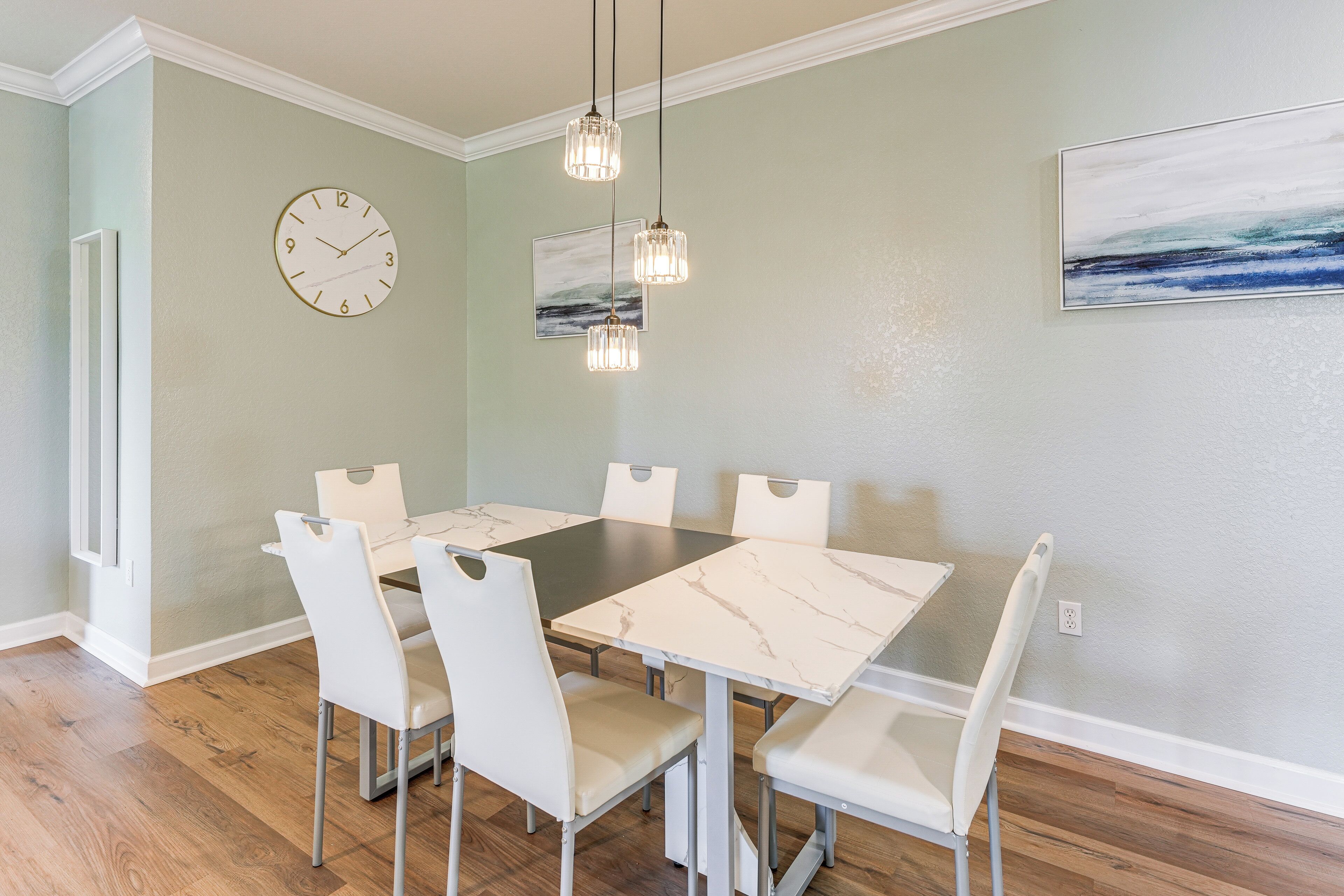 A dining area with a marble table, white chairs, pendant lights, a clock, and artwork on the wall.