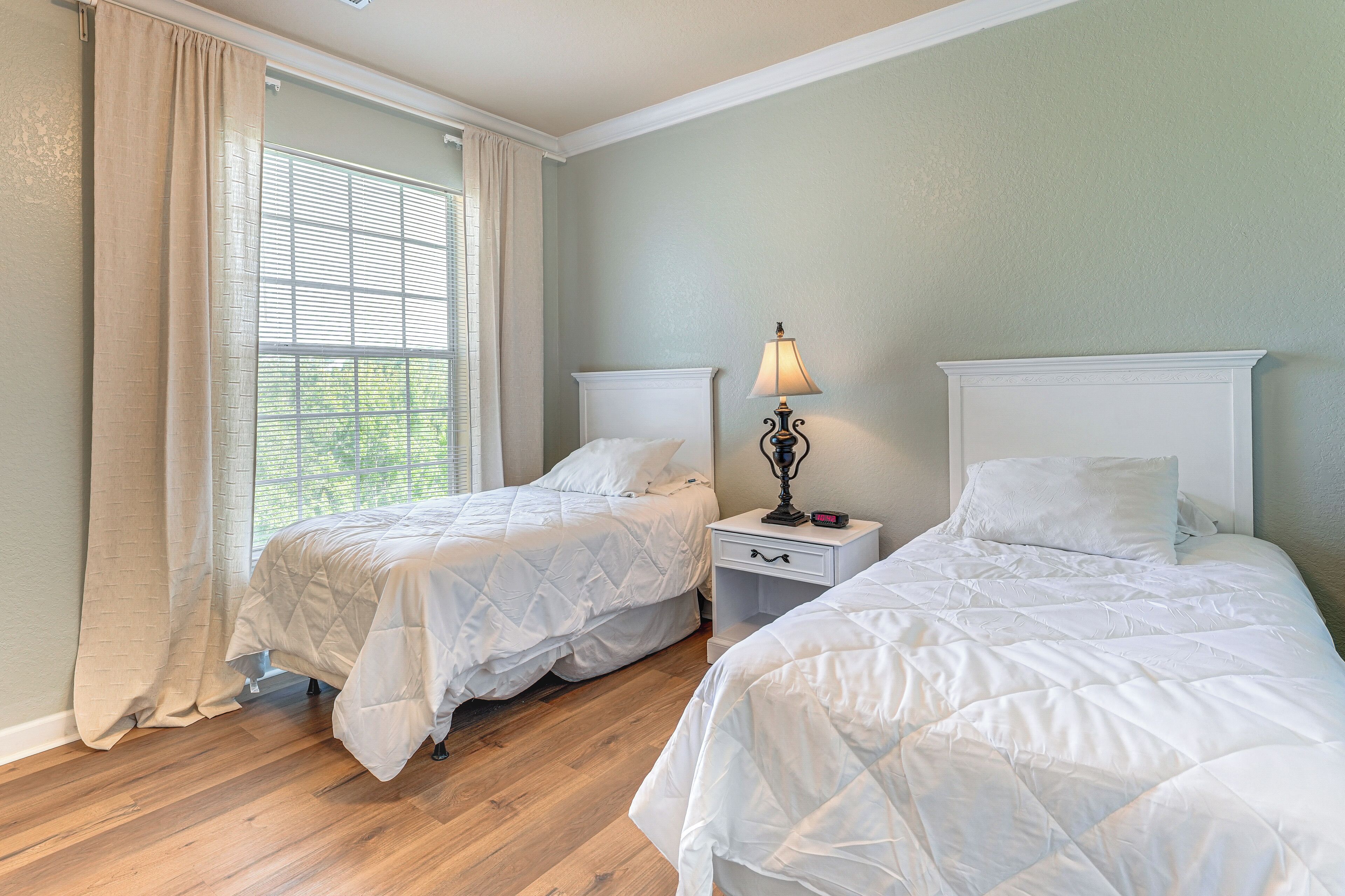 A room with two beds, a nightstand with a lamp, and a window with curtains.