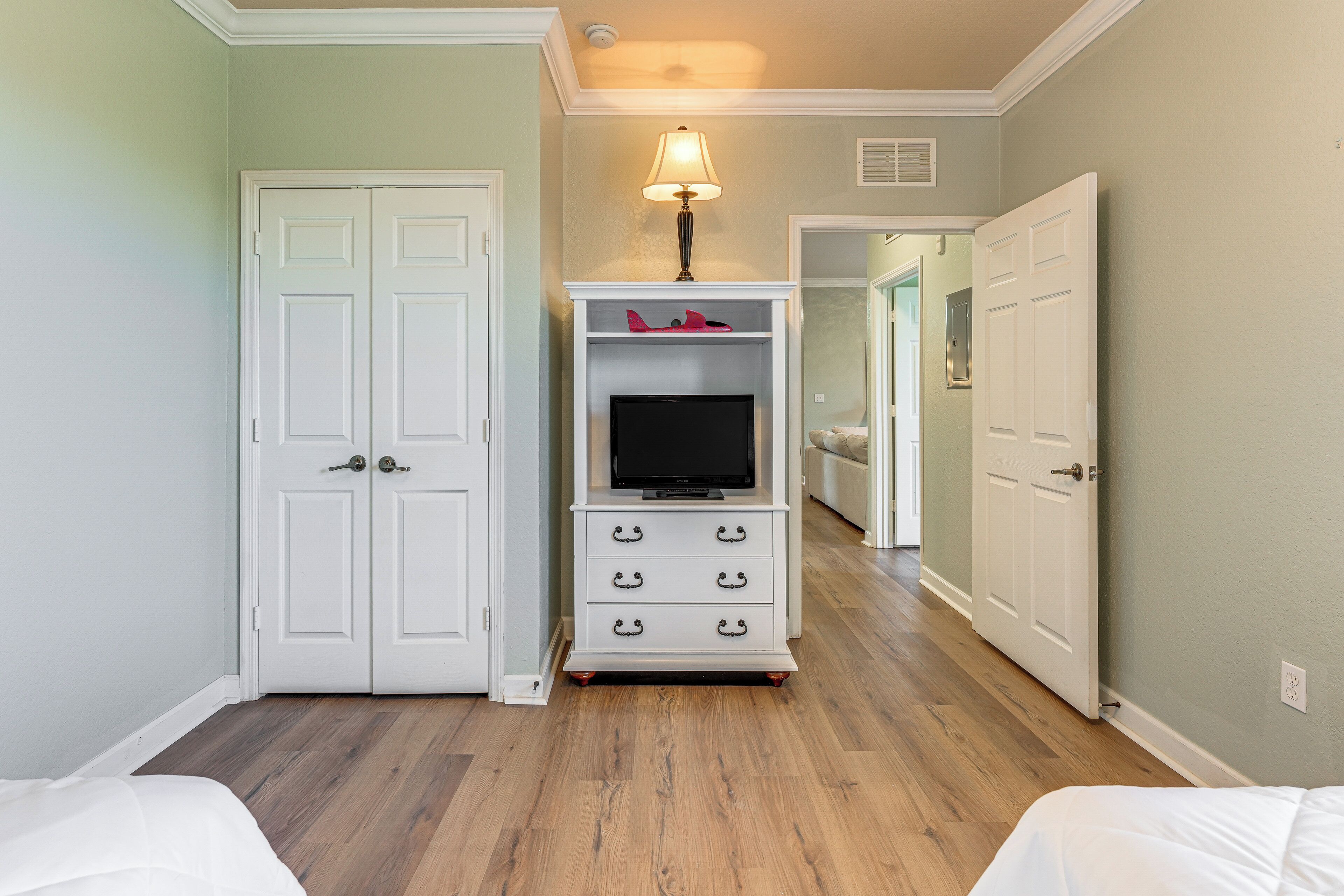 Une chambre d’hôtel avec un plancher en bois, des murs blancs et un téléviseur posé sur un meuble. On peut voir deux portes et un couloir.