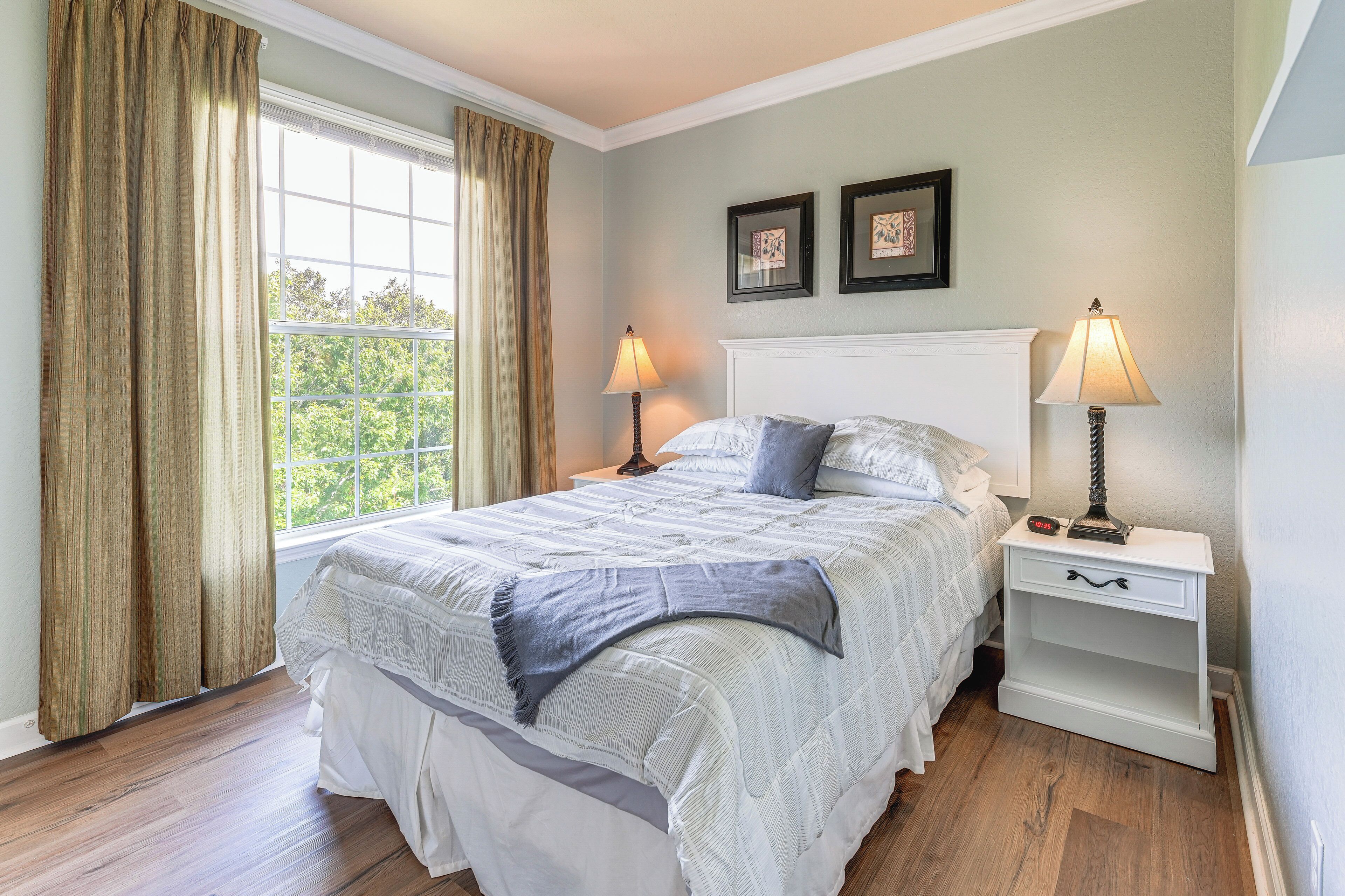 A neatly arranged bedroom with a large bed, two bedside tables with lamps, a window with curtains, and two framed pictures on the wall.