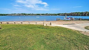 A lakeside view with a grassy area, a concrete path, and a dock with boats. - Cozy Mancelona Home: Ski, Golf & Snowmobile! (Mancelona)
