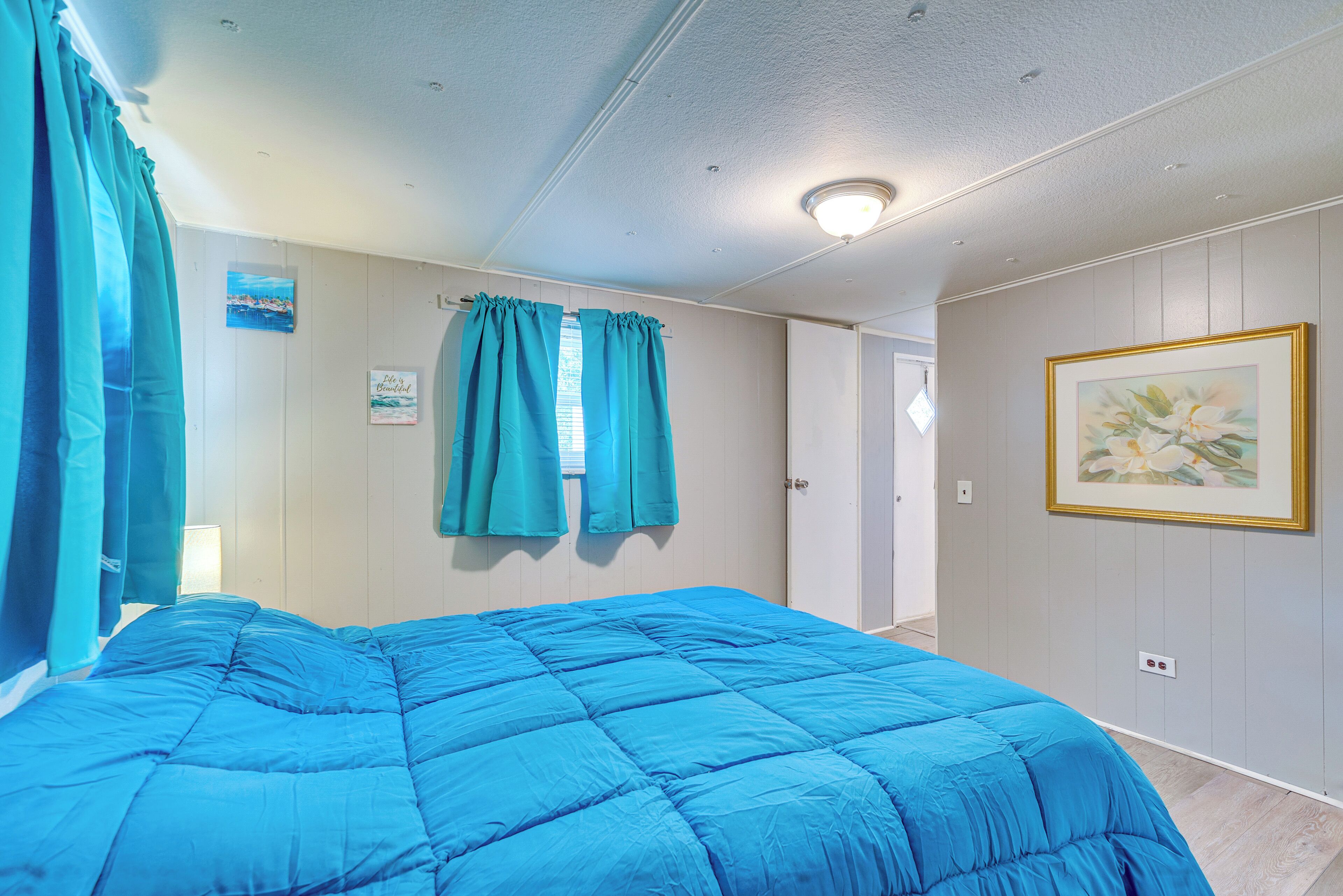 A bedroom with a large bed, teal curtains, a painting on the wall, and a door leading to another room.