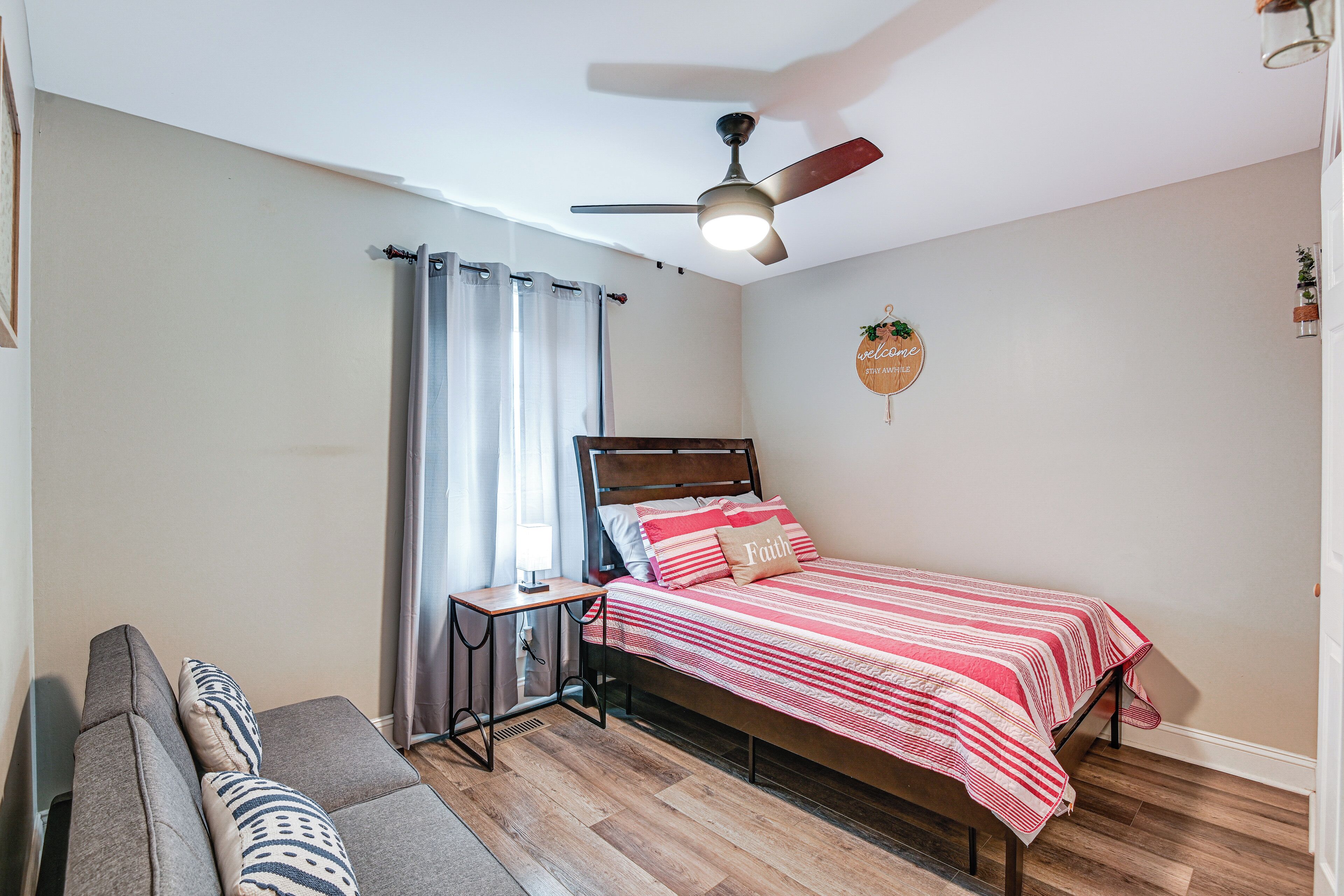 Una camera da letto con un letto, una ventola a soffitto, un comodino, una finestra con tende e una piccola pianta fissata al muro.