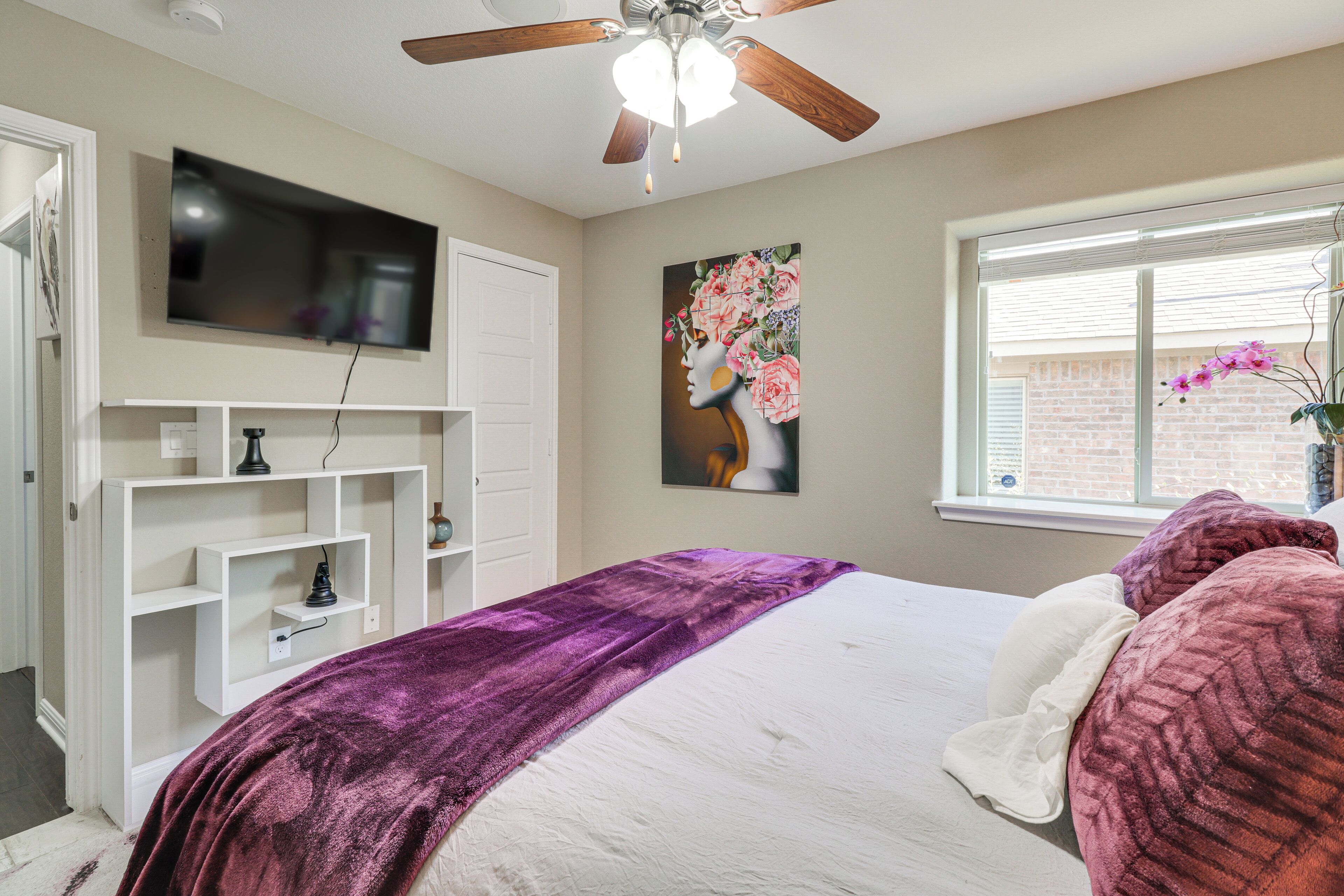 A bedroom with a bed, a ceiling fan, a TV mounted on the wall, and a window with blinds.