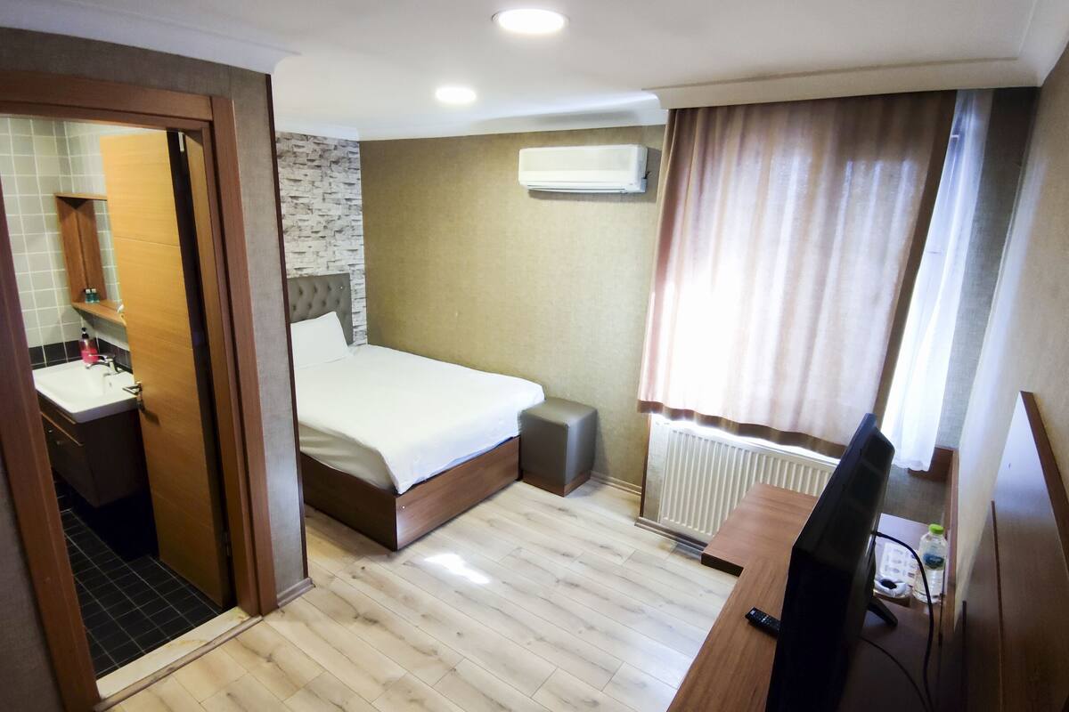 A hotel room with a bed, a desk with a TV, a bathroom with a sink, and a window with curtains.