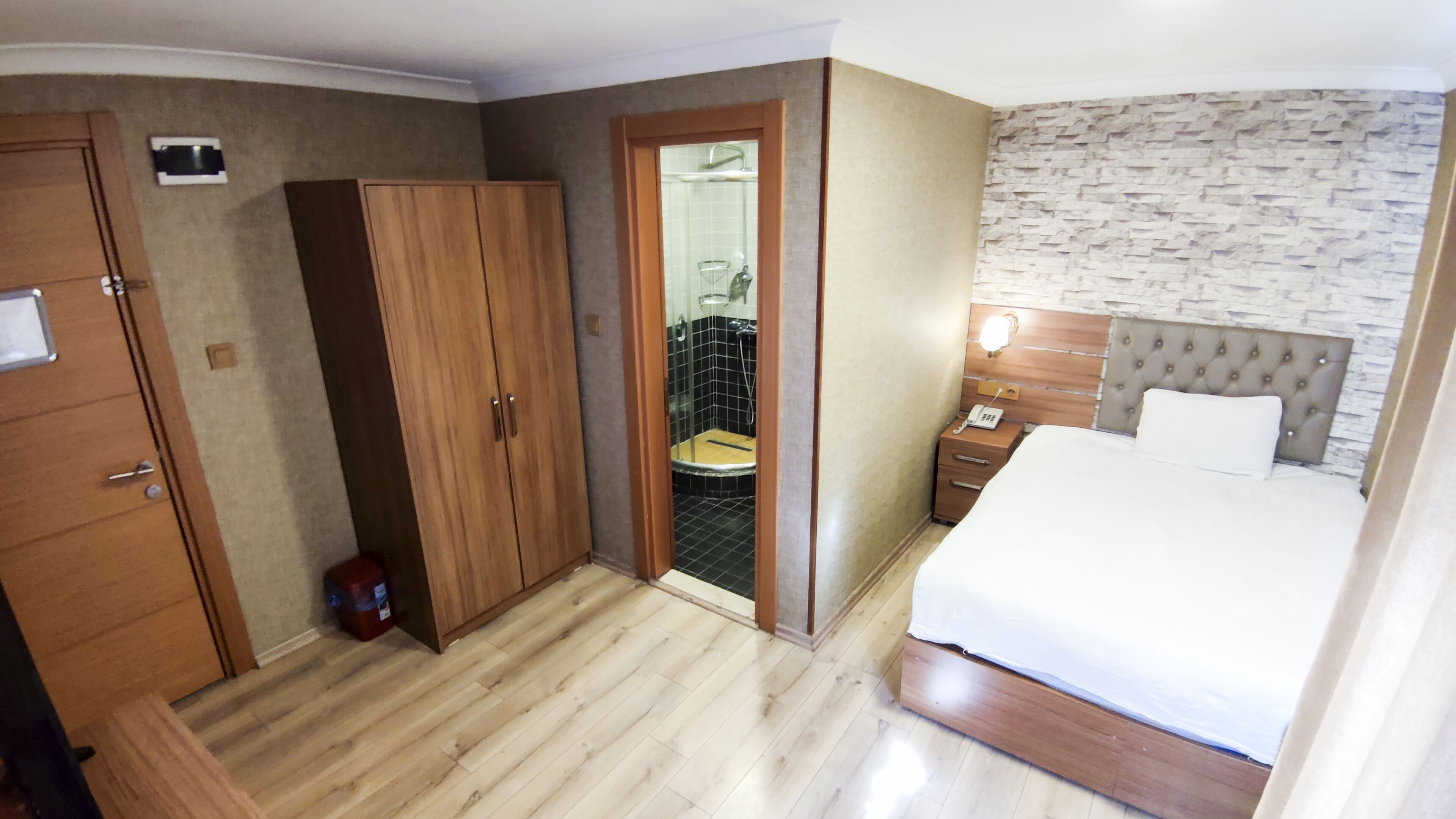 A hotel room with a bed, a wardrobe, a bathroom with a shower, and a small table with a phone.