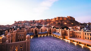 A rooftop terrace with intricate railings and seating areas, overlooking a historic fort and surrounding buildings.