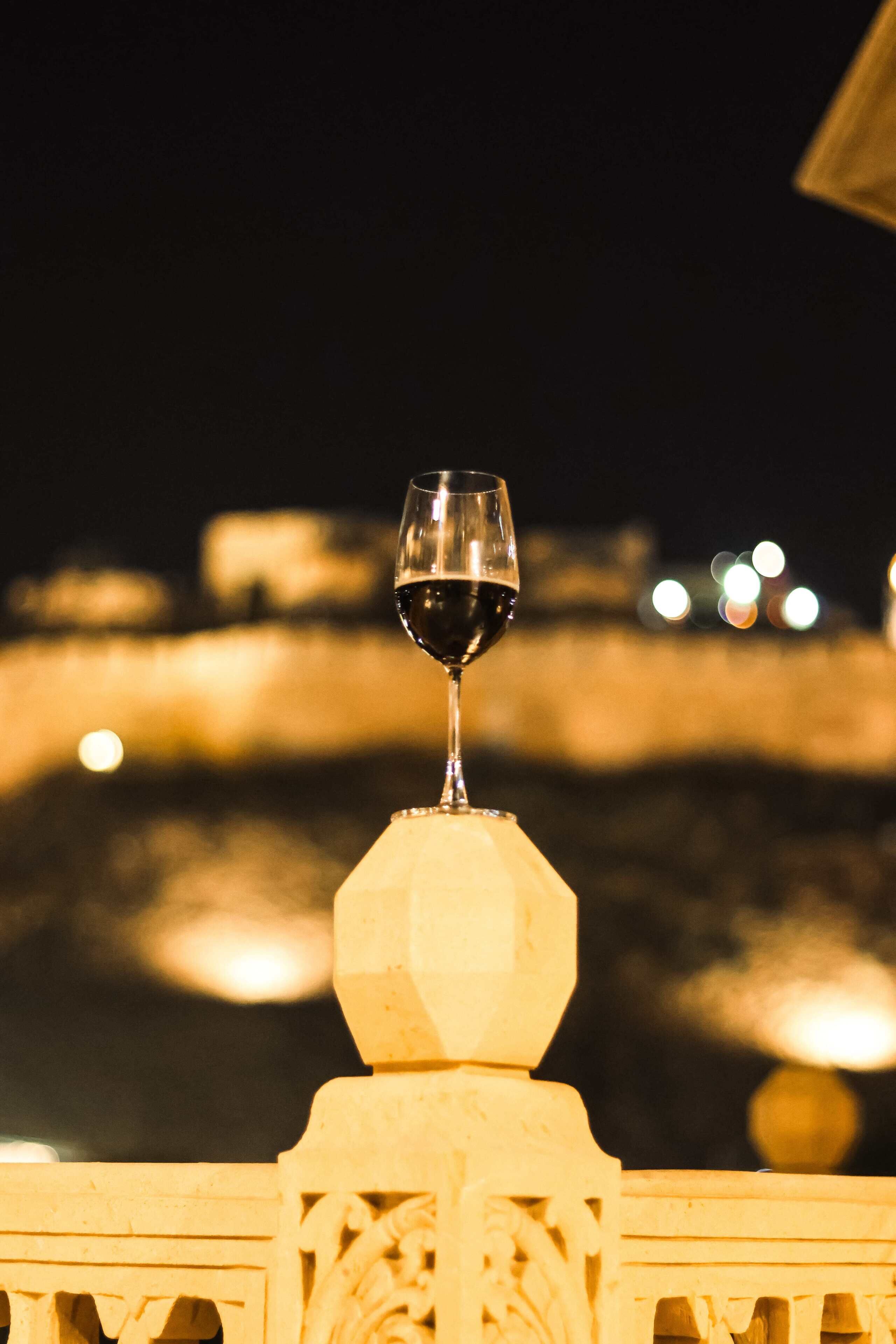 Een glas rode wijn op een balustrade met uitzicht op een wateroppervlak met lichtjes op de achtergrond.