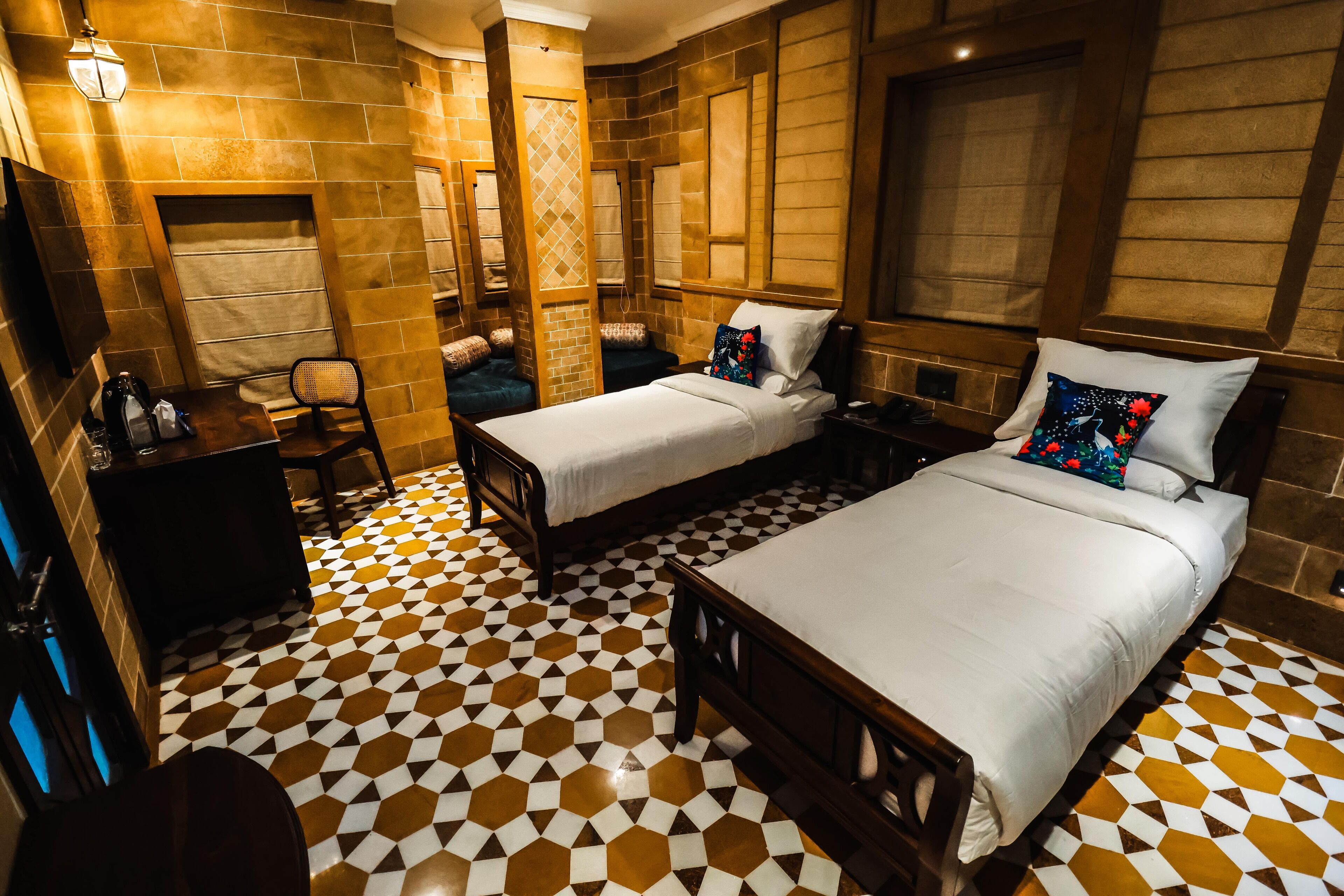 A hotel room with two beds, a desk, and a chair. The room has a checkered floor pattern and wooden paneling on the walls.