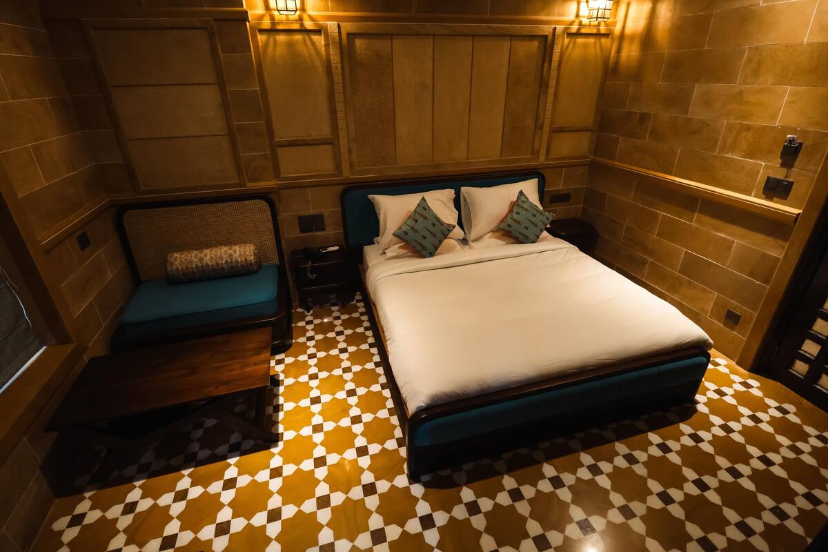 Ein Hotelzimmer mit Schachbrettmuster-Boden, einem Bett mit blauer Kopfteil sowie einer kleinen Sitzecke mit Kissen und Wandleuchten.