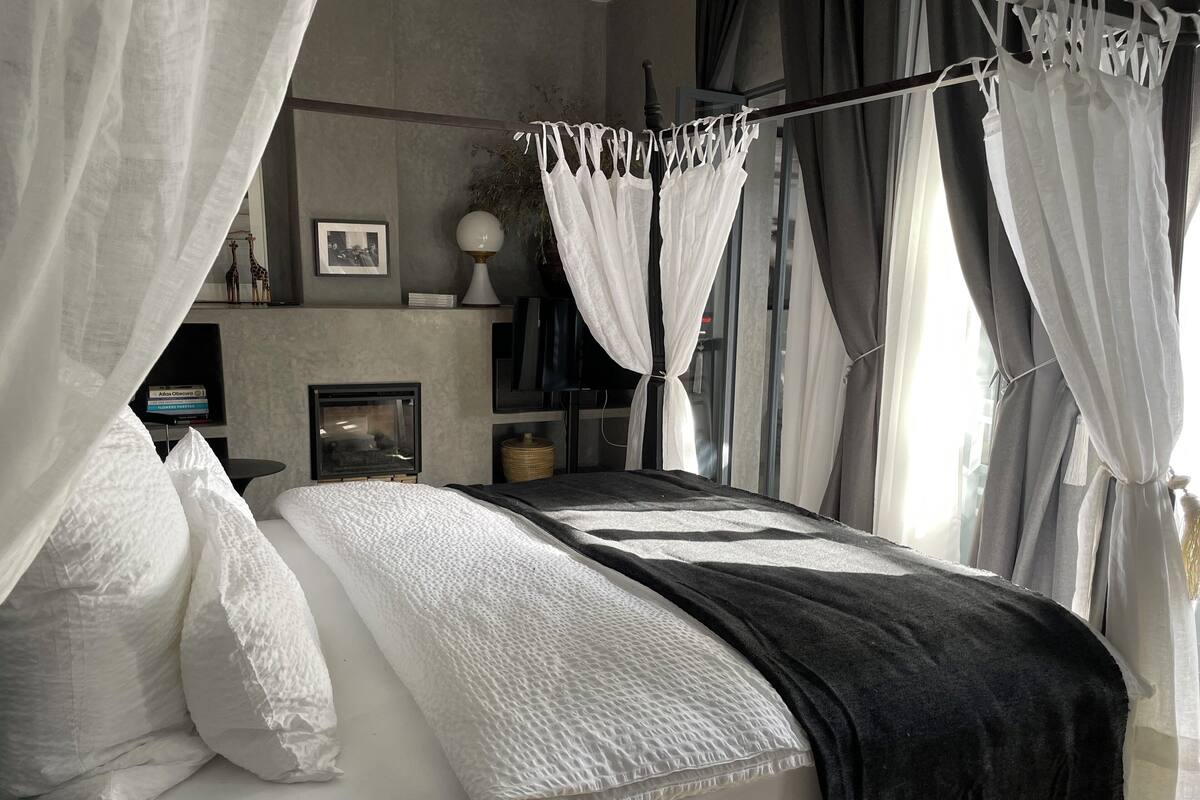 A bedroom with a bed, white bedding, a bedside table, a lamp, a framed picture, and a window with curtains.