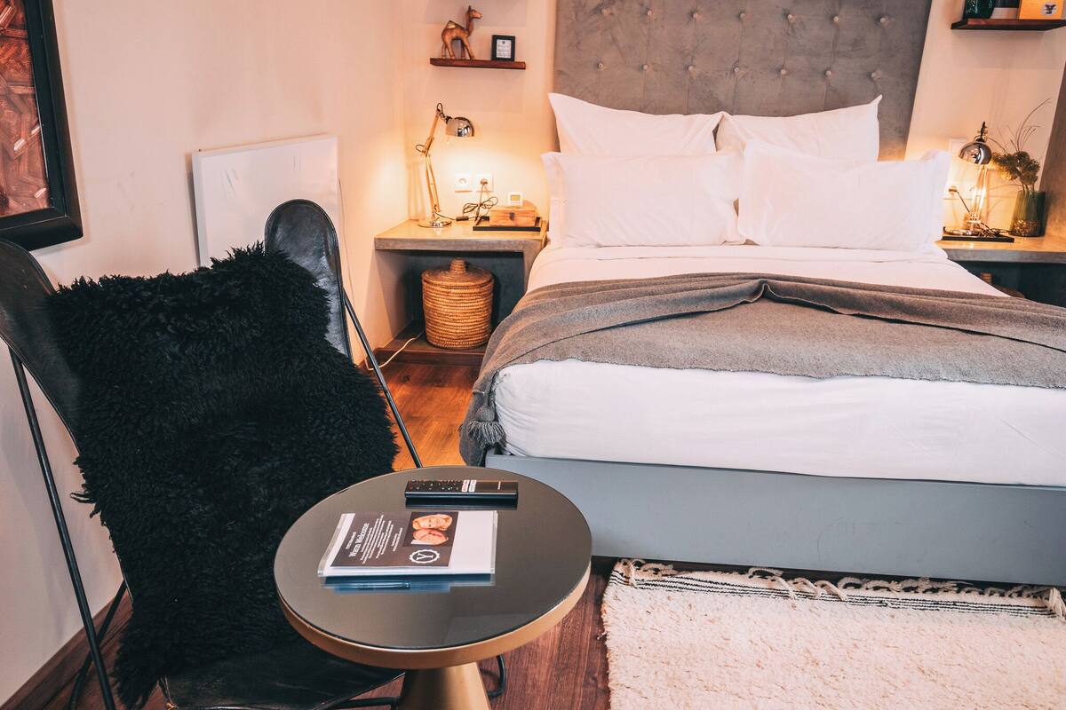 A neatly arranged bedroom with a bed, a chair, a small table with a book, and a bedside lamp.