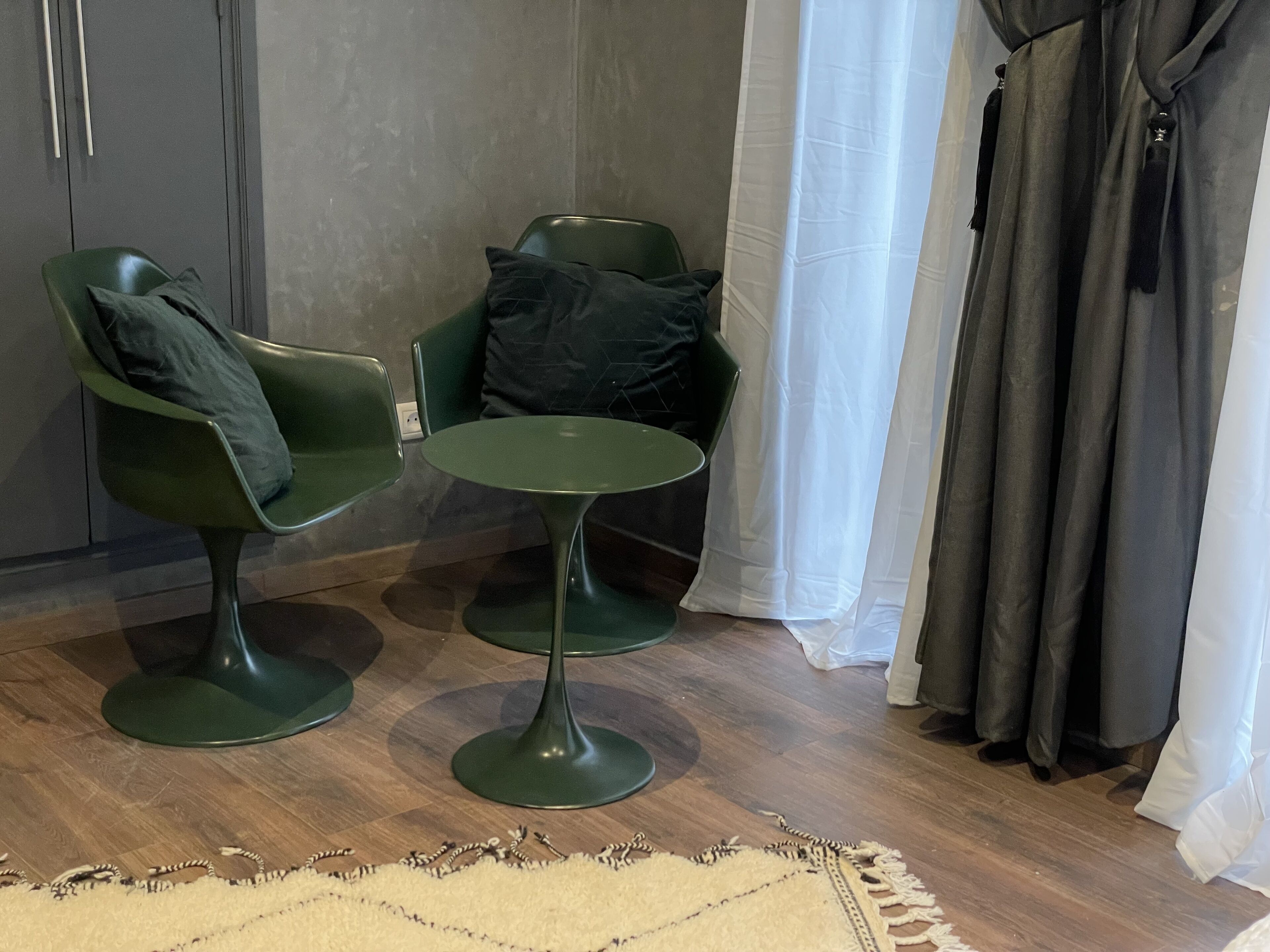 Een kamer met twee groene stoelen, een kleine ronde tafel, een houten vloer, een witte vloerkleed en een grijze muur met een deur.