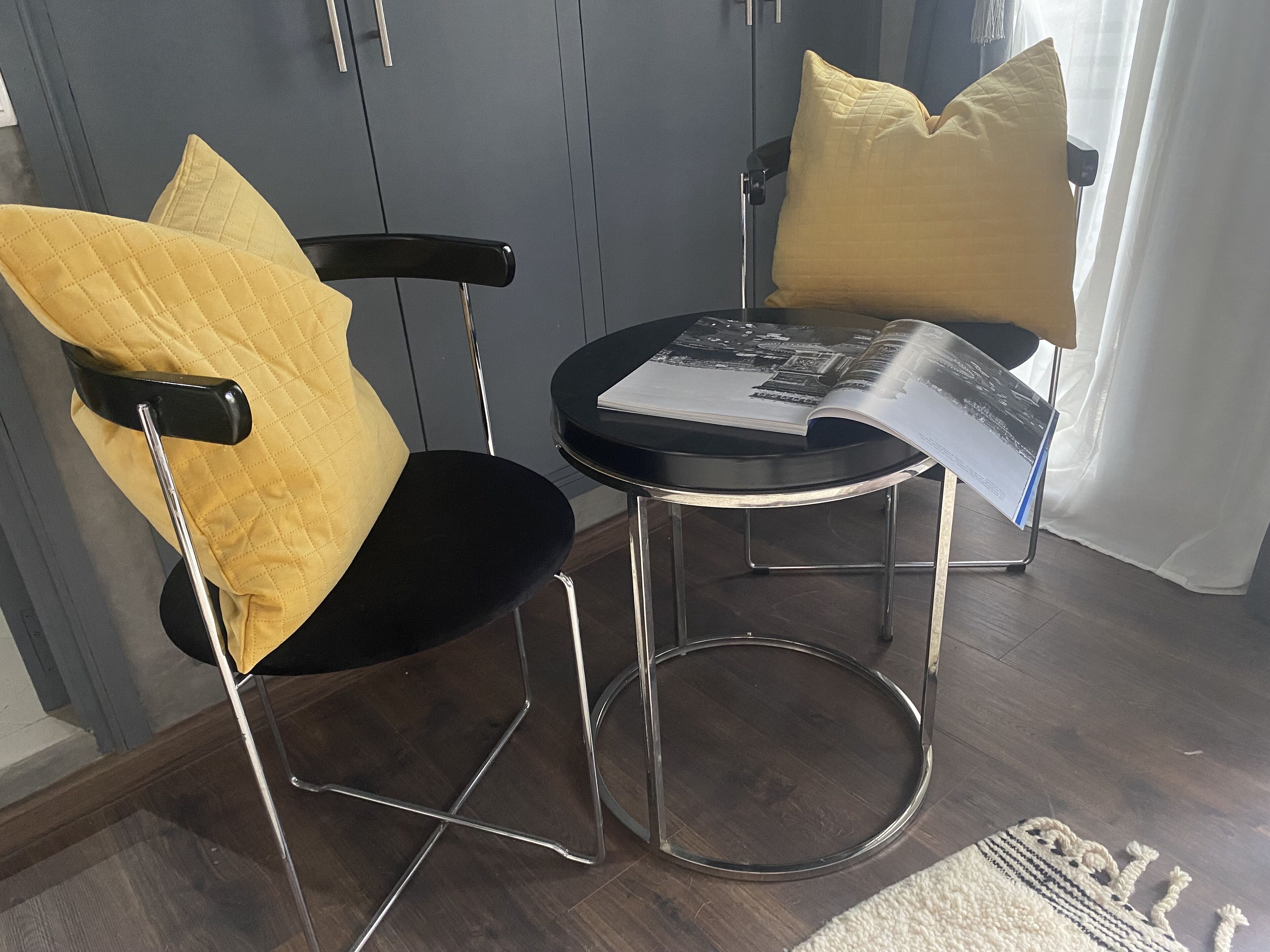 A chair with a black frame and a yellow cushion, a small round table with a magazine on it, and a white curtain in the background.