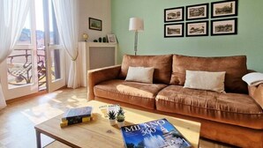 A cozy living room with a brown sofa, a wooden coffee table with books and a magazine, and a large window with curtains. - Little Broggi - 15mins walking from Central Station. (Milano)