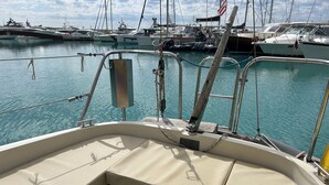 Het interieur van een boot met een zachte zithoek, metalen relingen en uitzicht op de jachthaven met andere boten.