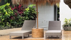 Two wicker lounge chairs with a woven table in between, set on a tiled patio with lush greenery and a thatched umbrella overhead. - Summer of Surf Hostel Uluwatu (Ungasan)
