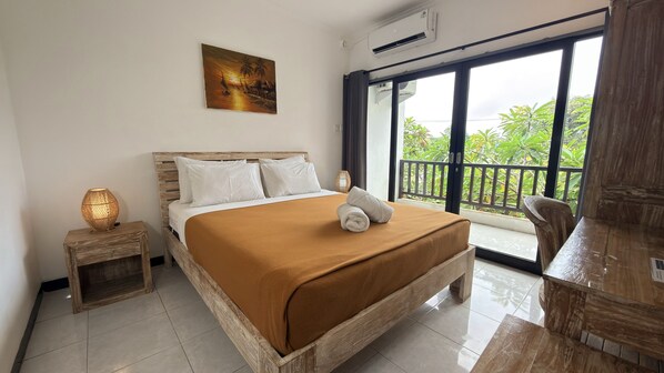 A bedroom with a bed, a nightstand, a painting, and a balcony with a view. - Summer of Surf Hostel Uluwatu (Ungasan)
