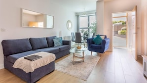 A modern living room with a dark blue sofa, a glass coffee table, and a blue armchair. There is a large window providing natural light and a view of the outside.
