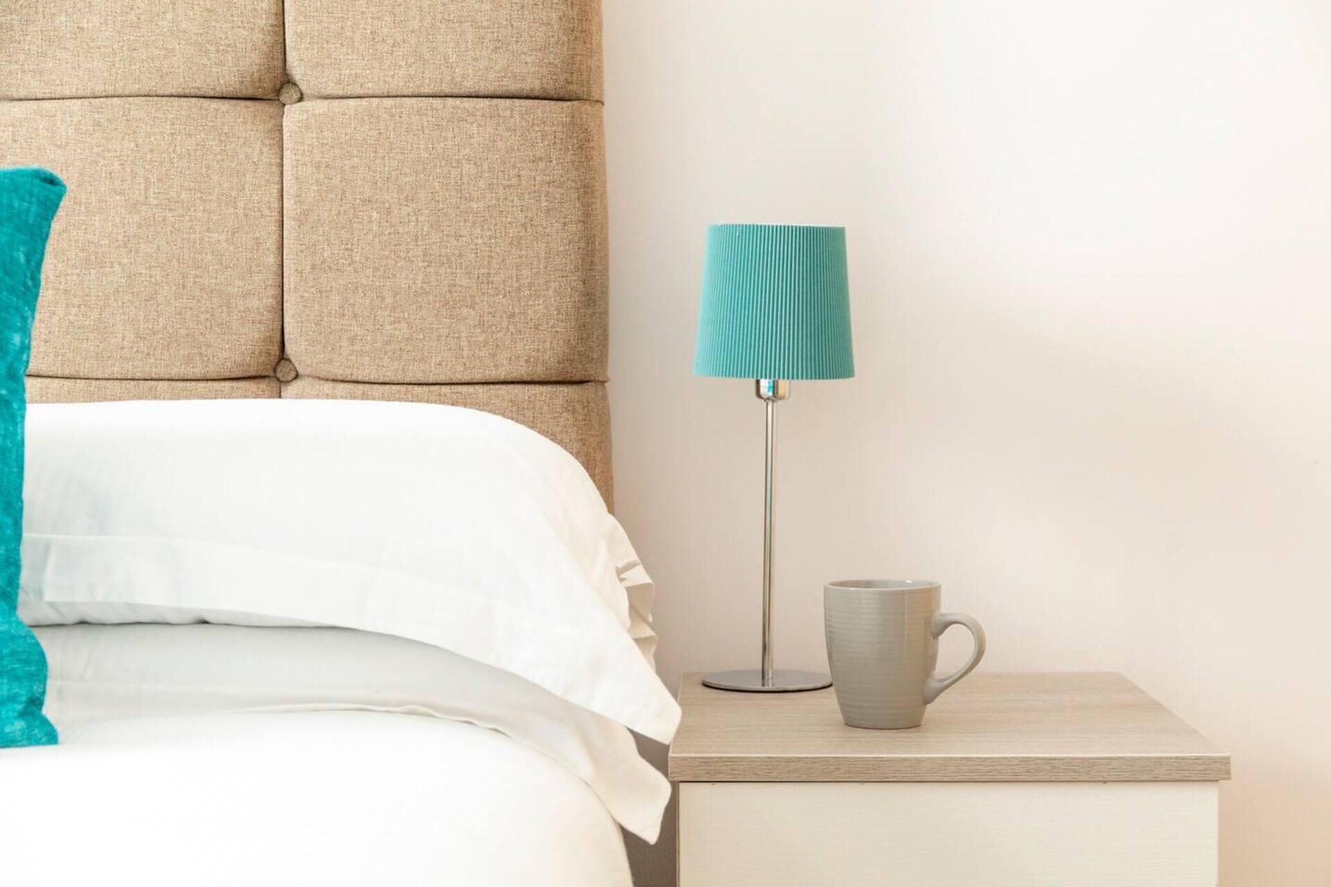 A neatly made bed with white linens, a teal throw pillow, a grey mug on a bedside table, and a tall floor lamp with a teal shade.