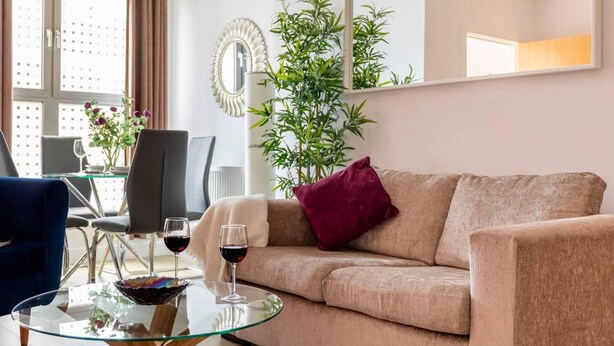 Ett vardagsrum med en beige soffa, ett soffbord i glas, två vinglas, en mörkblå fåtölj, ett matbord med stolar och en stor krukväxt.