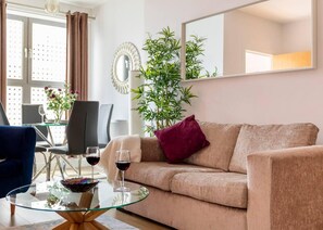A living room with a beige sofa, a glass coffee table, two wine glasses, a dark blue armchair, a dining table with chairs, and a large potted plant. - Castle Haven Apartment, Redhill (Redhill)