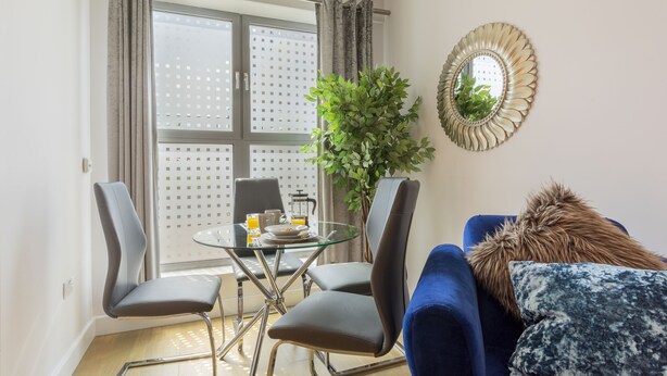 Een moderne woonkamer met een glazen eettafel, twee stoelen, een blauwe bank, een decoratieve spiegel en een potplant.