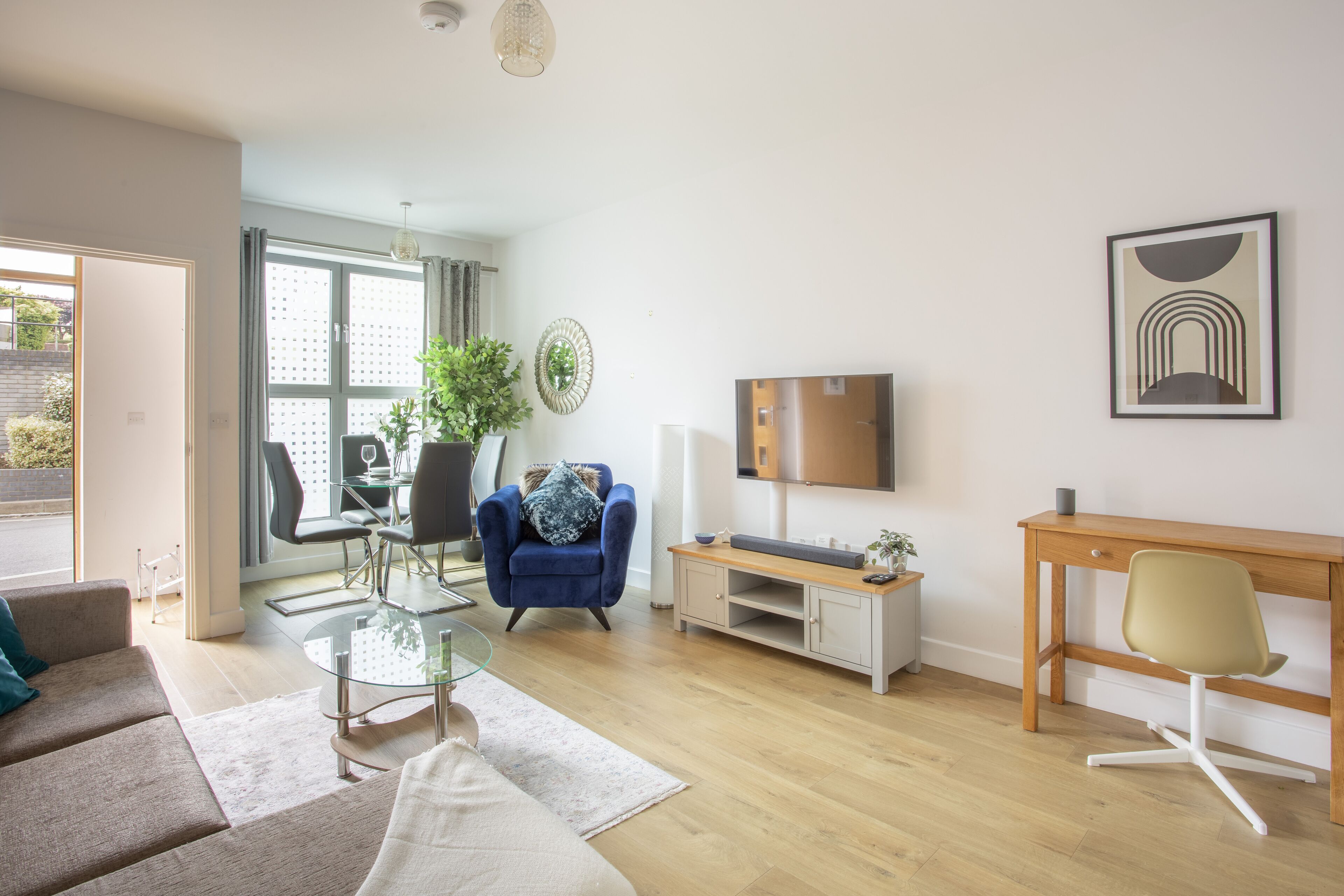 A modern living room with a sofa, a blue armchair, a glass coffee table, a wooden desk, and a TV.