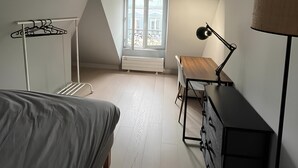 A bedroom with a sloped ceiling, a bed, a desk, a chair, a lamp, a radiator, and a window with a view of buildings. - Spacious Stunning 6th Floor Apartment in Renovated "Haussmannian" Building (Paris)