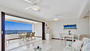 Amplia sala de estar con balcón que da al mar, con un sofá blanco, una mesa de comedor y ventiladores de techo.