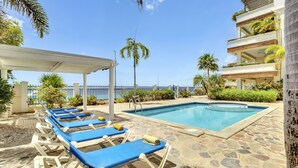 Zona de piscina con sillones de descanso y una piscina, rodeada de palmeras y un edificio con balcones.