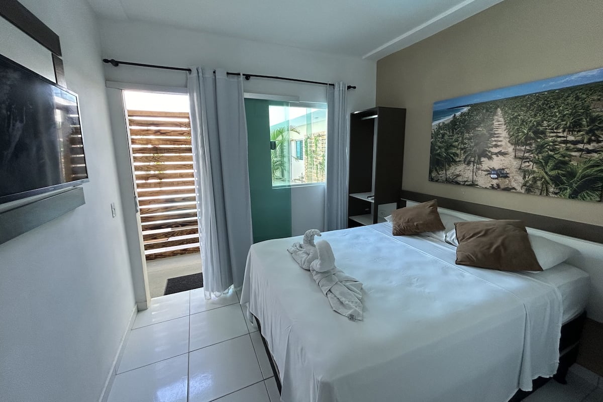 A hotel room with a bed, a TV, a painting, and a view of a building outside.