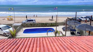 A beach view with a swimming pool, beach chairs, umbrellas, and cars parked near the shore. - Pousada Ondas do Mar (Ipojuca)