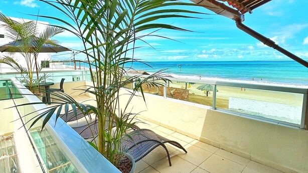 A balcony with a view of the beach, a palm tree, and a glass railing. - Pousada Ondas do Mar (Ipojuca)