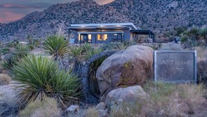 A modern house with a flat roof and large windows, surrounded by desert vegetation and a mountain backdrop. - Sunset Heights By Homestead Modern (Joshua Tree)