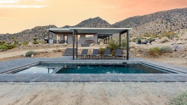 A modern outdoor pool area with lounge chairs and a covered seating area, surrounded by desert landscape and mountains. - Sunset Heights By Homestead Modern (Joshua Tree)