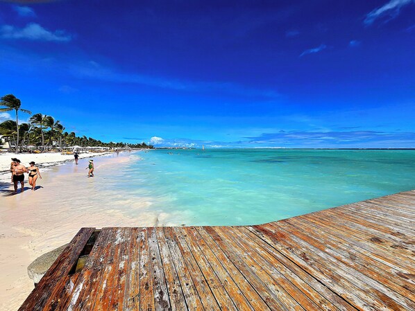 A wooden pier leading to a beach with clear turquoise water, palm trees, and a distant lighthouse. - Ocean Front Marina Sub Penthouse (Punta Cana)