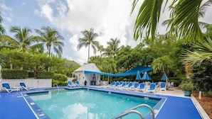 A swimming pool with blue deck and white lounge chairs, surrounded by palm trees and a small building. - THE PELICAN PEARL ~ Charming 2 BR/1 Ba Condo in Truman Annex! (Key West)