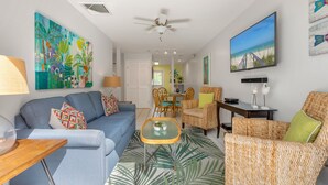 A modern living room with a blue sofa, wicker chairs, a glass coffee table, and a ceiling fan. - THE PELICAN PEARL ~ Charming 2 BR/1 Ba Condo in Truman Annex! (Key West)