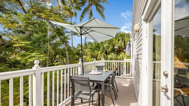 A covered outdoor dining area with a table and chairs, surrounded by a white railing and lush greenery. - THE PELICAN PEARL ~ Charming 2 BR/1 Ba Condo in Truman Annex! (Key West)
