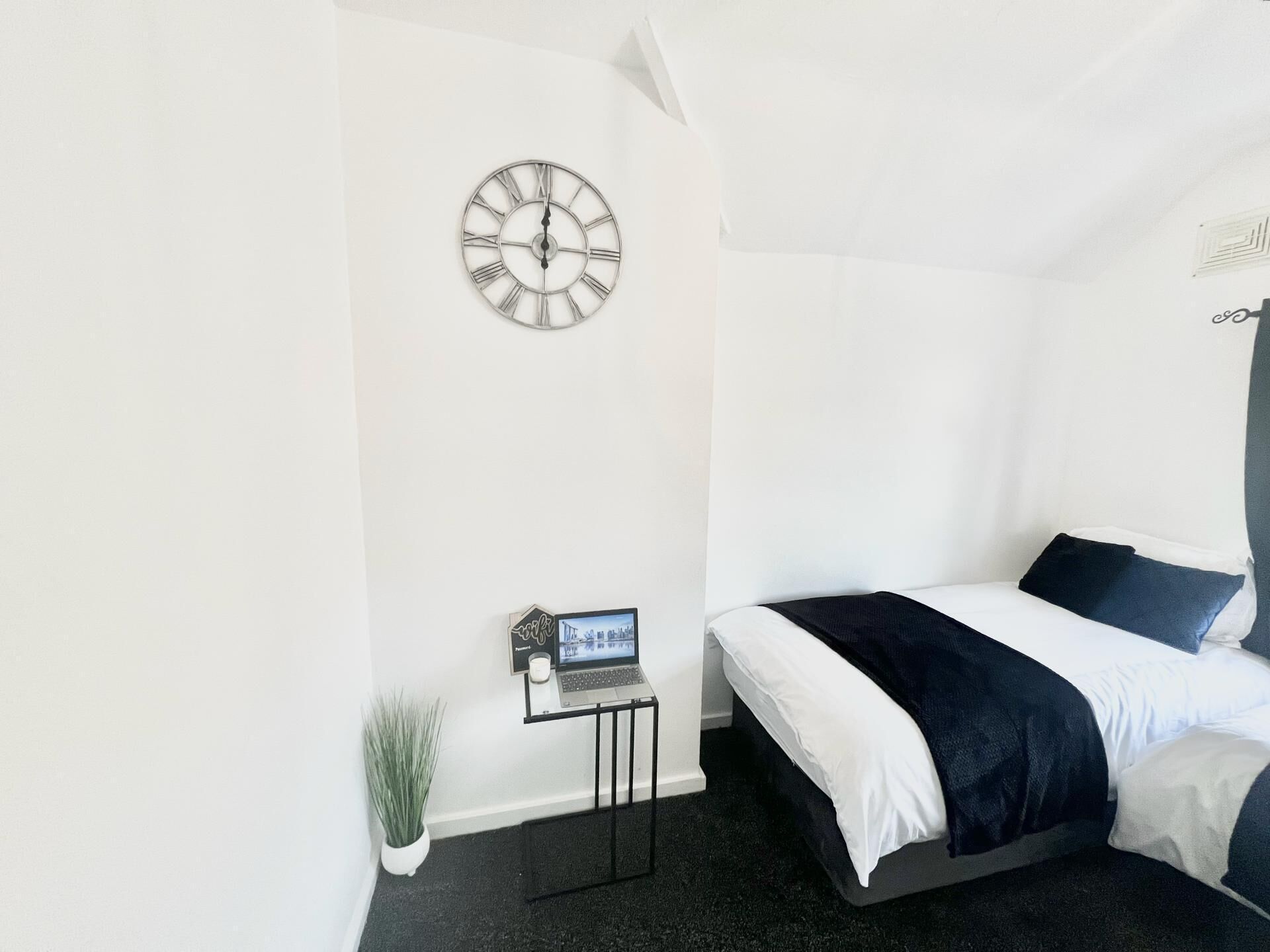 Ein Schlafzimmer mit einem Bett, einer Wanduhr, einem Laptop auf einem kleinen Tisch und einem Blumentopf.